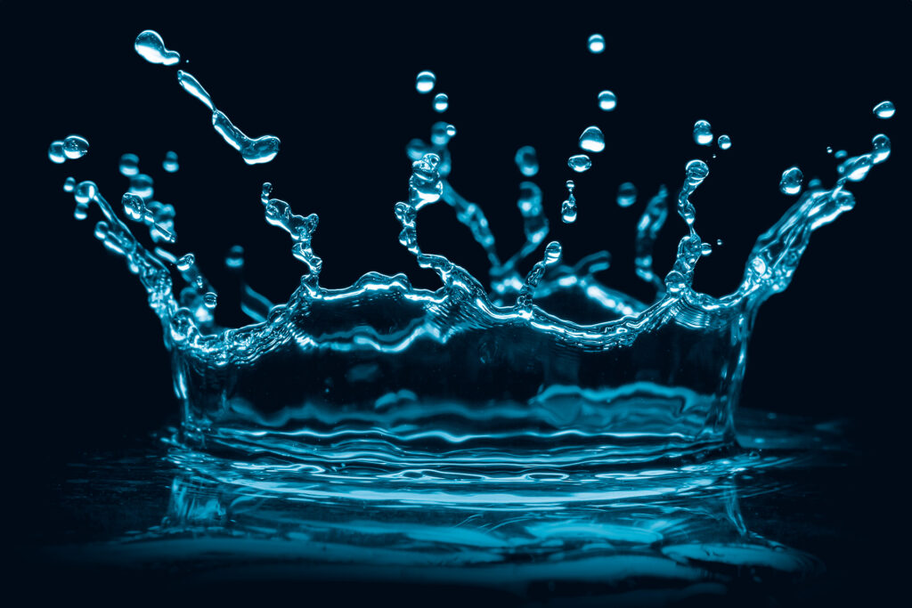 a blue-tinted freeze-frame image of a water droplet disrupting a smooth surface of water and creating a crown-shaped splash